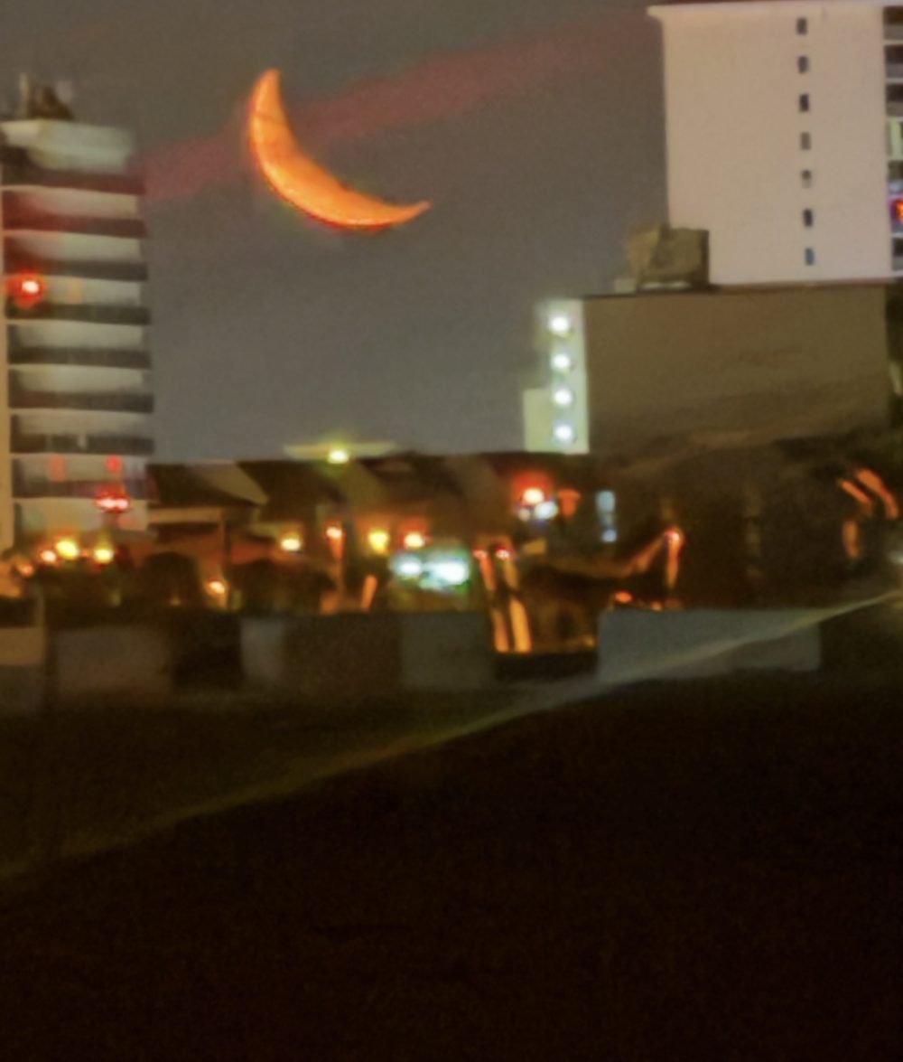 An orange crescent moon low over beachfront highrises